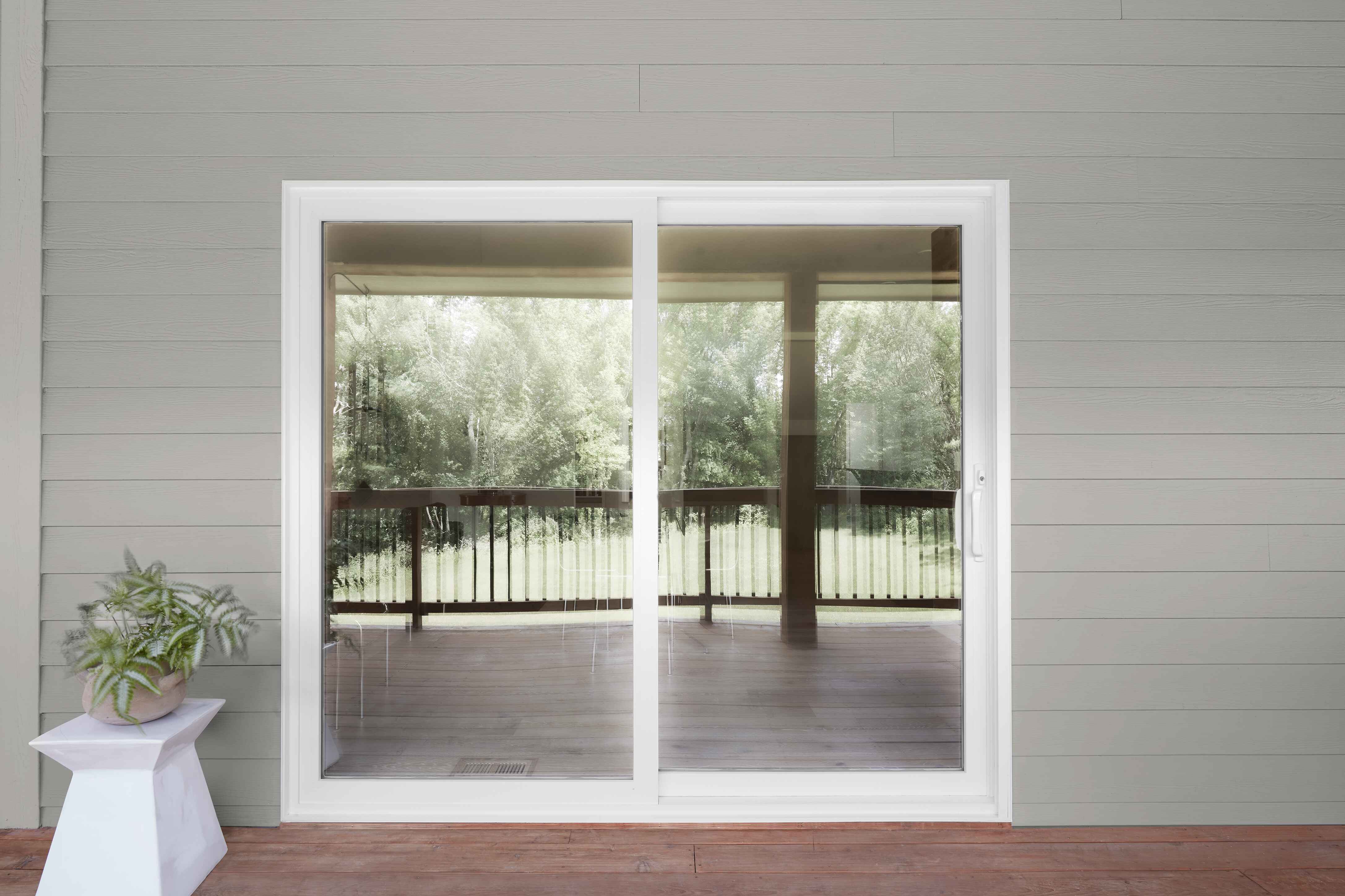 White sliding glass door reflecting a wooden deck and green trees outside, with a potted plant on a white stand to the left.