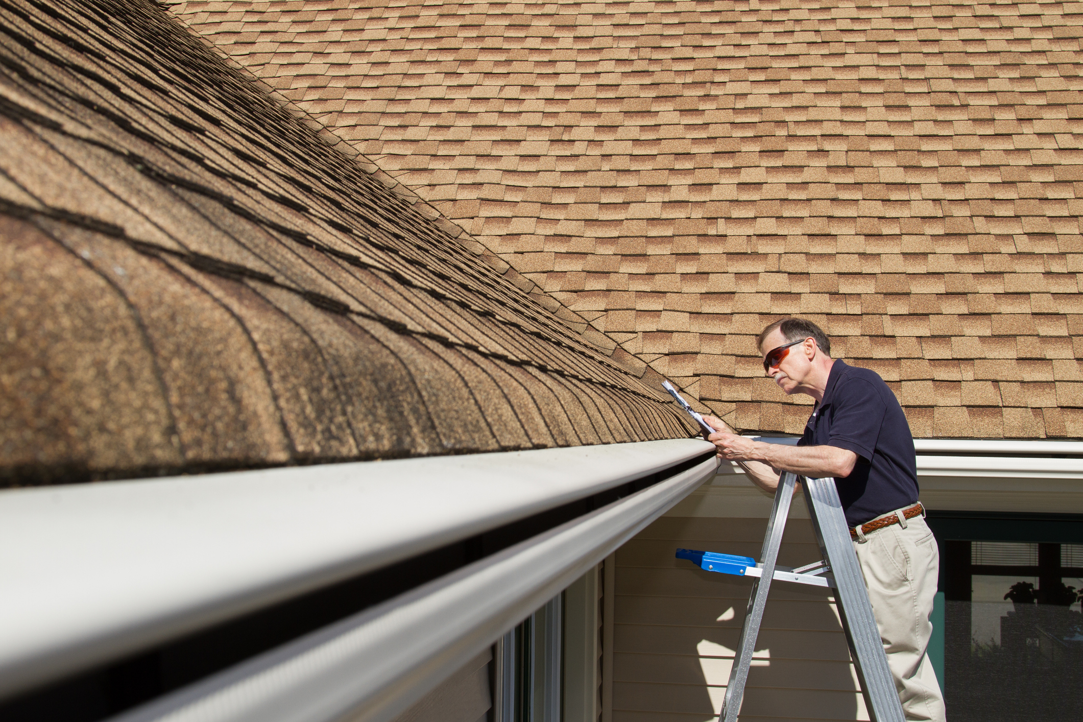 A man on a ladder inspecting gutters and shingles along the roofline of a home.